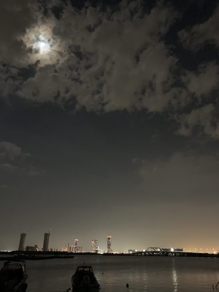 夜空に浮かぶ月と雲、対岸のビル群の光が水面に反射する静かな夜景