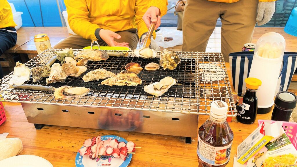 卓上コンロで牡蠣や貝類を焼く牡蠣小屋での食事風景、テーブルには調味料や飲み物も並ぶ