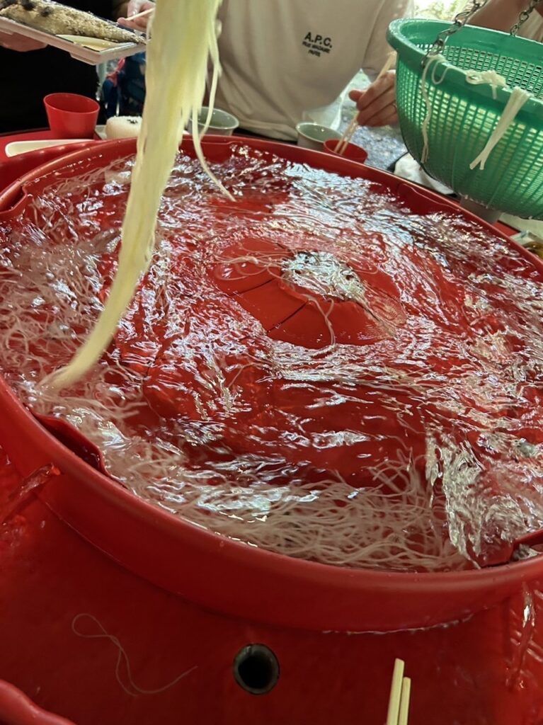 回転式の赤いそうめん流し機で冷たいそうめんを楽しむ様子。緑のざると食事中の人の手元も見える
