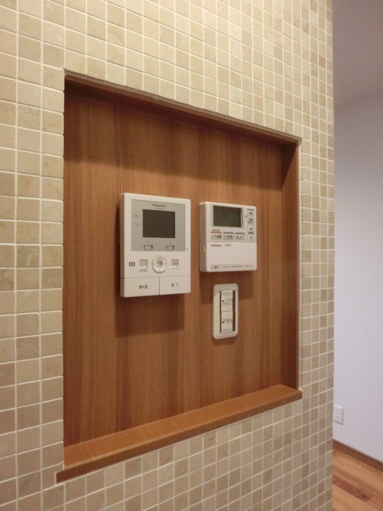 モザイクタイル壁に埋め込まれたスイッチニッチ。木目の背面にリモコンやスイッチがすっきりと収まっている