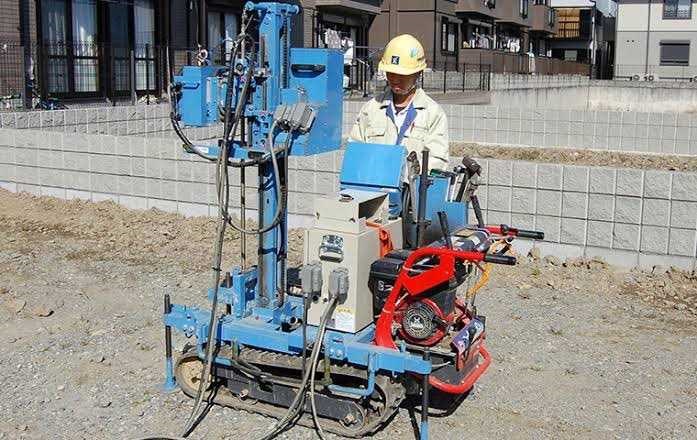地盤調査用の機械を操作する作業員。住宅地の一角で、青いボーリングマシンを使って地盤の強度を確認している様子